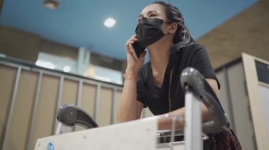 Havaalanında siyah koruyucu yüz maskesi ile telefon konuşması yapan genç bir kadın bagaj arabası, Corona virüsü salgını, yeni normal sosyal mesafeler...