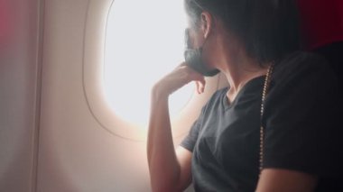 Uçakta pencere kenarında oturan genç Asyalı kadın yolcu, bilet kartı, covid-19 yeni normal seyahat sosyal mesafeyi koru, yolcu biletini kontrol et, salgın tehlikesi seyahat riski