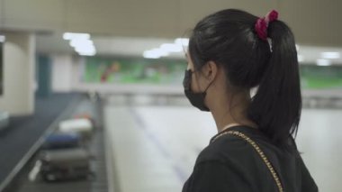 COVID-19 salgını. Asyalı kadın bavulunu valiz teslim salonunda beklerken siyah koruyucu maske takıyor. Havaalanı varış salonunda kemerini takıyor, yeni normal bir hayat sürüyor. Yalnız seyahat eden bir kadın, toplu taşıma riski taşıyor.