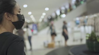 COVID-19 korona virüsü salgını. Asyalı genç bir kadın siyah yüz maskesi takıyor. Havaalanındaki bagaj teslim yerinde bagaj bekliyor. Yeni normal bir hayat, toplu taşıma, insanlar seyahat etmeye başlıyor. 