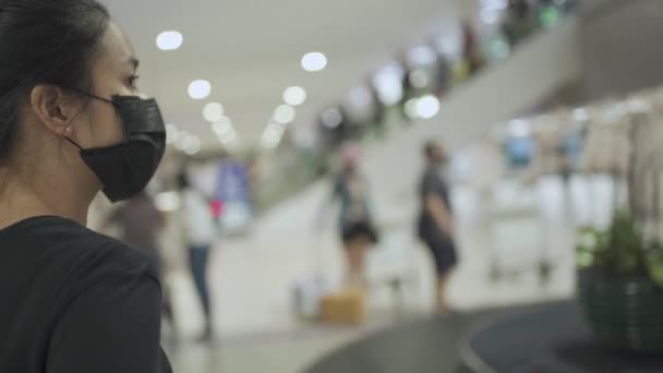 Covid コロナウイルスパンデミック アジアの若い女性は 空港ターミナル内の手荷物クレームエリアで荷物を待って黒いフェイスマスクを着用します新しい通常の生活 公共交通機関 人々は旅行し始めます — ストック動画