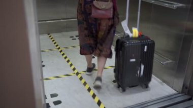 Asyalı kadın yüz maskesi takıyor. Havaalanı terminalinde asansörden in, seyahat bagajı, covid-19 salgını, yere yeni normal sosyal mesafe işareti. Uzak dur.