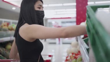 Yüz maskesi takan bağımsız bir Asyalı kadın raftan erzak seçiyor, market rafının önünde durup perakende süpermarketin içindeki ürünlere bakıyor, covid19 Corona virüsü salgını, yiyecek seçiyor.