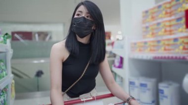 Asyalı genç kadın siyah koruyucu yüz maskesi market koridorunda alışveriş arabalarını itiyor, kadın kilitlenirken tedarik alıyor, tüketim ve ekonomik etki Coperid-19 yeni normal bütçe yaşam süresi