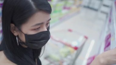 Asyalı bir kadın siyah maske takıyor raflardan erzak seçiyor, market raflarının önünde duruyor perakende süpermarkette ürünlere bakıyor, covid19 Corona virüsü salgını sırasında yaşam tarzına bakıyor.