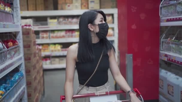 迷人的亚洲女人带着防护面罩在杂货店推着购物车 货物制造厂仓库得到供应 人类消费 衣食住行新常态 预算生活费用 超级市场走道 — 图库视频影像