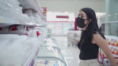 Genç Asyalı bir bayan siyah maske takıyor. Perakende satış mağazalarında ürün seçiyor, raflarda ürünler yığılıyor, Corona virüsü salgını, bütçede yaşayan tüketici alışkanlığı satışlar, ev aletleri...