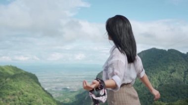 Dağın tepesinde duran Asyalı genç bayanın ağır çekimde kollarını uzat. Rüzgarın esintisini hisset. Dağlara ve bulutların arka planına bak.