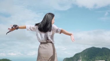 Dağın tepesinde duran Asyalı genç bayanın ağır çekimde kollarını uzat. Rüzgarın esintisini hisset. Dağlara ve bulutların arka planına bak.