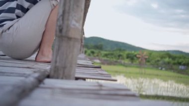 Genç bir kadın, eski tarım ahşap köprüsünde bacakları sallanarak oturuyor, Asya geleneksel pirinç tarlası çiftliği, sakin bir rahatlama, doğası güzel, yoğun şehirden kaçıyor, alt vücut parçalarından kaçıyor.