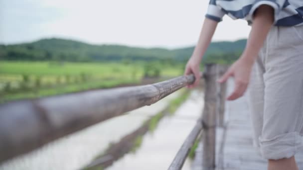 若い女性は木製の橋の手の鉄道 旅行の旅で彼女の腕を休める アジアの田舎側の村 水田の背景をぼかす 穏やかな風光明媚な非都市の澄んだ天気の空 人間の自由 — ストック動画