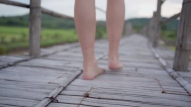 Eskimiş ahşap köprüde çıplak ayakla yürüyen açık tenli kadın, geleneksel Asya yapısı, yeşil pirinç tarlası ve arka planda dağ, kentsel olmayan manzara yerel doğaya dokunur, tasasız özgürlük