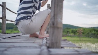 Ağır çekimde genç bir kadın, tahta köprünün kenarına oturmuş, bacaklarını sallıyor, Asya kırsal kesiminde geleneksel pirinç yetiştirme alanı, sakin rahatlama, alt vücut kesimi.