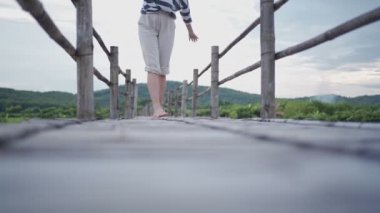 Genç bir kadın tahta köprüde çıplak ayakla yürüyor, eğlence bakımı ücretsiz mutlu seyahat, arka planda yeşil dağ, rahatlık doğada hayat, alçak görüş, kameraya doğru yürüme, yavaş çekim