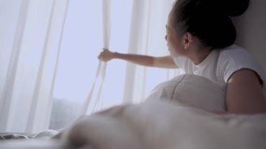 Genç Asyalı kadın uyanıyor ve perdeleri açıyor sıcak sabah güneşinin tadını çıkarmak için, pencere perdelerini açıyor ve pencereden dışarı bakıyor, yeni bir başlangıç, yeni bir gün başlıyor, güneş ışığı pencereyi aydınlatıyor