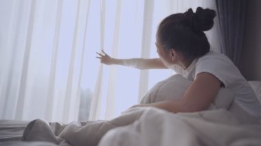 Genç Asyalı kadın uyanıyor ve perdeleri açıyor sıcak sabah güneşinin tadını çıkarmak için, pencere perdelerini açıyor ve pencereden dışarı bakıyor, yeni bir başlangıç, yeni bir gün başlıyor, güneş ışığı pencereyi aydınlatıyor