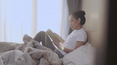 Genç Asyalı kadın rahat bir şekilde yatağın üzerinde uzanıyor kitap okuyor, verimli bir sabah aktivitesi, rahat yatak odası çarşafları gün ışığı, kişisel eğitim, yaratıcı bir an.