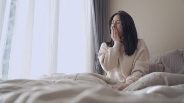 Genç, yetişkin Asyalı bir kadın uyanıp kolunu yatakta uzatıyor. Tembel bir sabah geç saatlere kadar uyanıyor. Rahat yatak odası perdeleri, doğal gün ışığı, kentsel yaşam tarzı. 