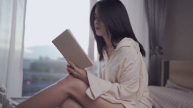 Genç Asyalı bayan yatakta yalnız başına kitap okuyor, ev ödevi sayfaları çeviriyor, ders notlarını gözden geçiriyor, ders bilgisi, gün ışığı, yatak odası penceresi ışığı, kişisel eğitim, final sınavı için ezberleyin. 