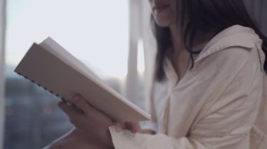 Genç, Asyalı bir kadın kitap okuyor. Yatakta oturuyor, kalın bir kazak giyiyor, sırtına battaniye örtüsü örtüsü örtmüş, hafta sonları sabah boş vakit geçiriyor, yatak odasının penceresinden gün ışığı görünüyor.