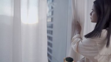 Genç Asyalı kadın sabahın erken saatlerinde uyanır açık pencere perdeleri, kahve fincanını yudumlar, bir fincan kahvenin tadını çıkarırlar ve yüksek katlı apartman dairesine, şehir manzarasına bakarlar.