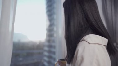 Genç Asyalı kadın yataktan kalktıktan sonra sabah kahvesini yudumlarken bir fincan kahvenin keyfini çıkarıyor ve pencereden dışarı bakıyor şehir manzaralı bir apartman dairesinde, sabahın erken saatlerinde kaygısızca dinleniyor.