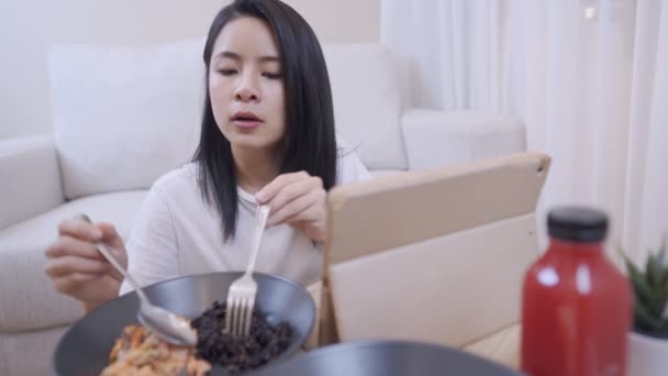 スプーンとフォークで食べる若いアジアの女性は 家のリビングルームで自分自身を楽しんで 背後にあるソファと座って シンプルなライフスタイル 一人で家に滞在 肯定的な女性 健康的な家庭料理ディナー — ストック動画