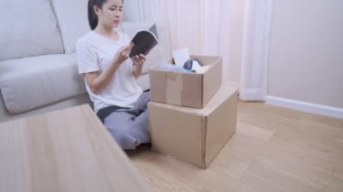 Asyalı güzel bir kadın yeni daireye bir şeyler ayarlamak için paket kutusunu açıyor, koltuğa oturuyor, birden fazla iş yükleme sahili, konsept içinde taşınma, evde kalma, orta çekim.
