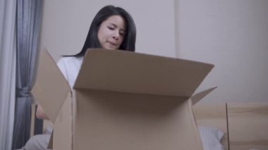 Genç Asyalı kadın yatak odasında oturuyor kahverengi paket kutusunu açıyor, kadın kutunun içinde eski eşyaları buluyor ne olduğunu merak ediyor, depo yöneticiliği yapıyor, taşınıyor, evde kalıyor, teslimat kutusunu açıyor.