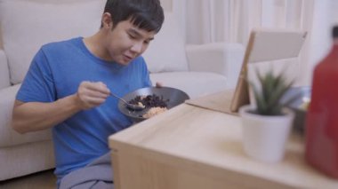 Genç Asyalı bir adam evde otururken kaşıkla yemek yiyor, basit bir yaşam tarzı, evde yalnız başına, olumlu duygular içinde, tablet, kablosuz teknoloji izlemekten zevk alıyor.