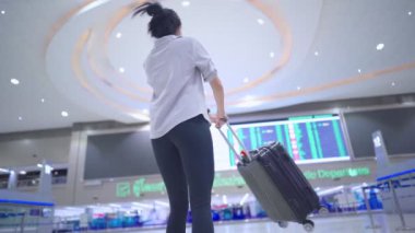 Genç bir kadın tıbbi maske takıyor, havaalanının zaman çizelgesine bakıyor, içi boş havaalanı terminalinde, covid-19 salgını sırasında seyahat riski, yeni normal sosyal mesafe, virüs yayılan önleme