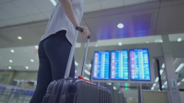 Genç bayanın vücudunun alt kısmı ayakta duruyor, seyahat valizli uçuş çizelgesine bakın, havaalanı terminalinde, hareket halinde, uçak biletleri kontrol servisi, valizli kadın.