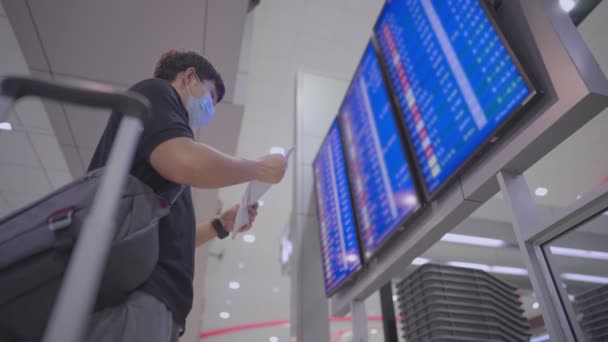 亚洲年轻男子戴口罩站在机场航班时刻表屏幕前 登机柜台区凭订票 在机场候机楼 男子和行李箱行李低角度向上看 — 图库视频影像