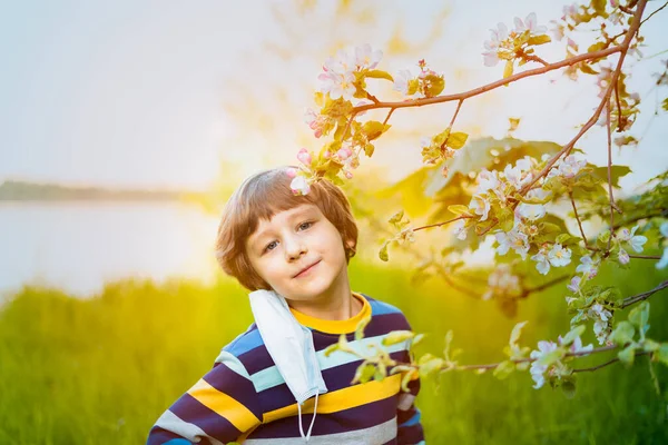 Cute boy take off medical face mask in flowering apple trees tying to smell flowers and looking at camera.. Happy caucasian child in park at sunset. Quarantine exit concept .