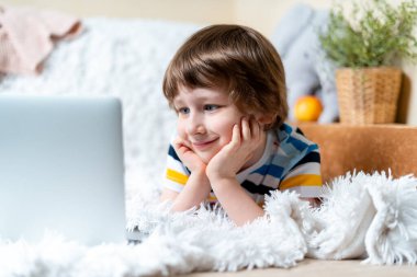 Uzaktan öğrenme çevrimiçi eğitim. Kafkasyalı gülümseyen çocuk evde tablet dizüstü bilgisayarla ders çalışıyor ve okul ödevi yapıyor. Kanepe ve battaniyeye uzanıp bilgisayara bakarken 