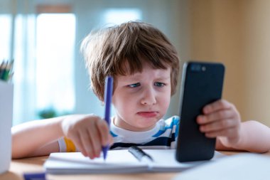 Uzaktan öğrenme çevrimiçi eğitim. Kafkasyalı üzgün çocuk evde akıllı telefonuyla ders çalışıyor ve okul ödevi yapıyor. Çocuk, Coronavirus karantinası nedeniyle cep telefonuyla masada oturuyor..