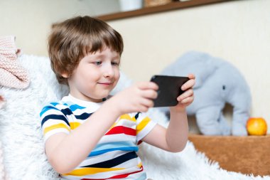 Mutlu küçük çocuk online oyun oynuyor, cep telefonuyla video izliyor, kanepede oturuyor ve oturma odasında eğleniyor. Komik mobil uygulamalar kullanarak gülümseyen küçük bir çocuk, evde boş vakitlerin tadını çıkarıyor.