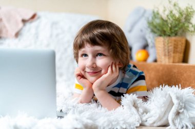 Uzaktan öğrenme çevrimiçi eğitim. Kafkasyalı gülümseyen çocuk evde tablet dizüstü bilgisayarla ders çalışıyor ve okul ödevi yapıyor. Yatağa uzanıp bilgisayara bakarken .