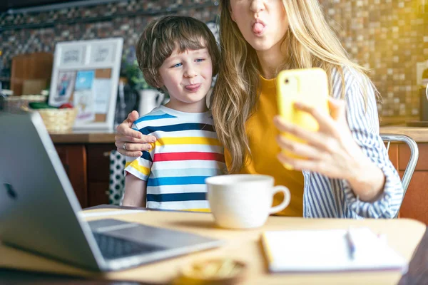 母亲和儿子露出舌头 让自己看着智能手机 一起在家里玩的开心在家里工作的妇女和带着电脑和电话的孩子上网接受教育的妇女 — 图库照片
