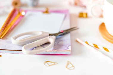 Masaüstü el yapımı DIY araçları. Beyaz çalışma masasının arka planında bir fincan kahve. Düz makas, kasetler, şakayıklar, altın kağıt ataçları, boş tebrik kartı, not defteri ve kalem..
