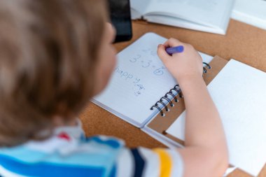 Okula dönelim. Uzaktan öğrenme çevrimiçi eğitim. Beyaz tenli gülümseyen çocuk evde akıllı telefonuyla ders çalışıyor ve okul ödevi yapıyor. Düşünen çocuk cep telefonuyla masada oturuyor..