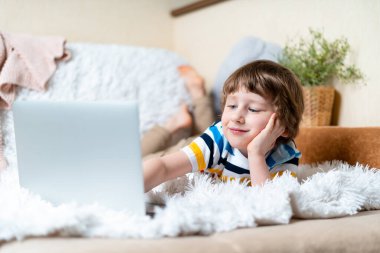 Okula dönelim. Uzaktan öğrenme çevrimiçi eğitim. Beyaz tebessümlü anaokulu çocuğu evde dizüstü bilgisayarla ders çalışıyor ve ödev yapıyor. Kanepe ve battaniyeye uzanmış bilgisayarla.