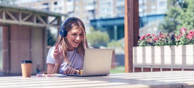Kulaklıklı bir kadın laptopuyla video görüşmesi yapıyor. Mutlu ve gülümseyen kız dışarıda çalışıyor ve kahve içiyor. Bilgisayar ve cep telefonu kullanarak. Uzaklık öğrenme çevrimiçi eğitim ve çalışma.