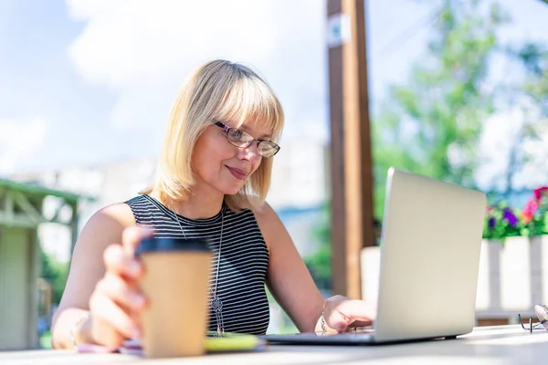 Vuxen kvinna i glasögon med videosamtal med bärbar dator utanför i parken. Glad och leende senior arbetar och dricker kaffe. Använda dator. Distansutbildning på nätet och nätbutiker — Stockfoto