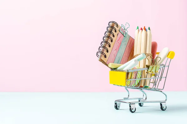 Buying school supplies, pencils, tape, notebook and crayons. Stationery for kids in a shopping cart on pink turquoise background, copy space. Back to school concept
