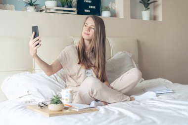 Mutlu ve gülümseyen neşeli kız akıllı telefon kullanıyor, yatakta selfie çekiyor ve kahve içiyor. Sarışın, cep telefonu ve görüntülü arama kullanıyor. Uzaklık öğrenme çevrimiçi eğitim ve çalışma