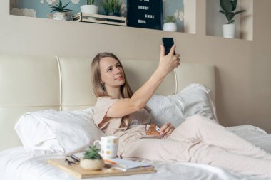 Mutlu ve gülümseyen neşeli kız akıllı telefon kullanıyor, yatakta selfie çekiyor ve kahve içiyor. Sarışın, cep telefonu ve görüntülü arama kullanıyor. Uzaklık öğrenme çevrimiçi eğitim ve çalışma