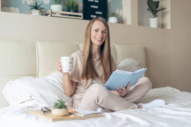 Mutlu ve gülümseyen, gevşemiş, gevşemiş, defter okuyan, evde çalışan, yatakta kahve içen bir kadın. Sarışın kız hafta sonunun keyfini çıkar ve kanepede aletlerle meşgul olarak dinlen.