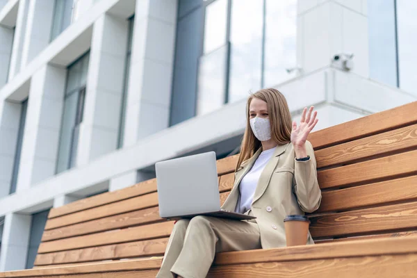 Beautiful business woman in medical protective mask and having a video call at laptop sit down on bench outside on a urban city street. Happy lady girl distance learning and online education.