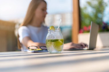Yazın güneşli bir şehirde cam kavanozlardan limonlu kokteyl içen güzel bir kız öğrenci. Kadın oturur ve dizüstü bilgisayarın başında yazar. Seçici odak.